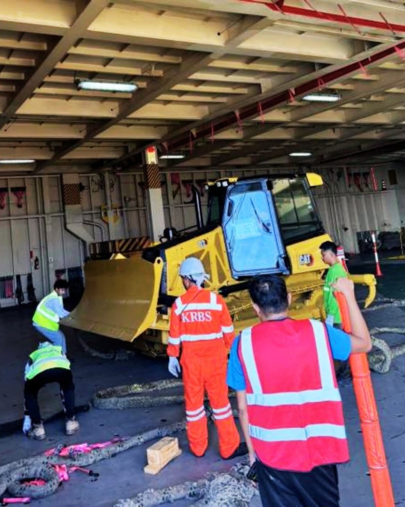 Workers securing yellow CAT dozer with ropes and chocks in RORO ship hold
